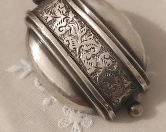 May include: A silver-toned antique compact mirror with an oval base and a decorative band featuring an engraved floral pattern. The band is centered on the mirror and has rounded edges. Small spherical accents are on each end of the band.