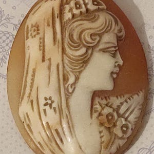 Pode incluir: Um broche de camafeu com o perfil de uma mulher com um véu e flores no cabelo. O camafeu é esculpido em uma concha marrom claro e engastado em uma moldura de metal dourado.