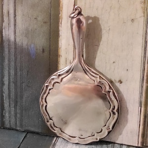 Puede incluir: Un espejo de mano plateado con un borde decorativo festoneado. El espejo tiene un mango largo y delgado con un lazo en la parte superior. La superficie del espejo refleja el entorno. El diseño general es vintage y elegante.