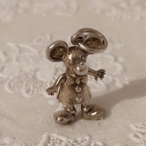 May include: A silver-toned mouse figurine with large, rounded ears and a bow tie. The mouse is wearing a dress with three buttons and has large, rounded feet. The figurine is standing with its arms outstretched.