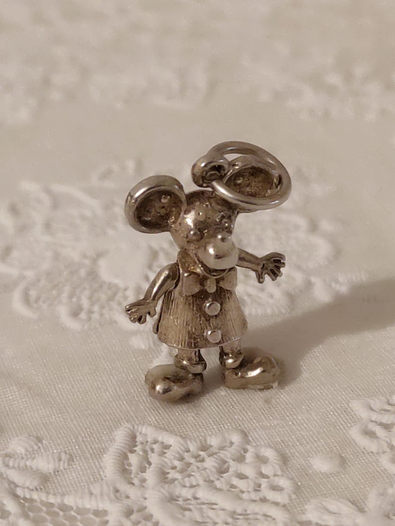 May include: A silver-toned mouse figurine with large, rounded ears and a bow tie. The mouse is wearing a dress with three buttons and has large, rounded feet. The figurine is standing with its arms outstretched.