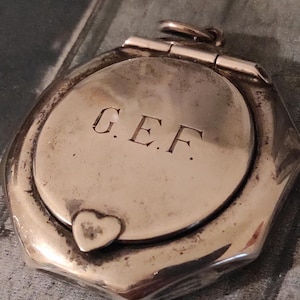 Puede incluir: Un relicario octogonal plateado con tapa abisagrada. El relicario presenta un centro circular con las iniciales grabadas "G.E.F." y un pequeño cierre en forma de corazón. El relicario tiene un lazo en la parte superior para una cadena.