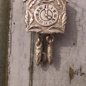 May include: Silver-toned cuckoo clock charm with a black clock face and Roman numerals. The charm has a small window with a bird inside and two dangling bells.