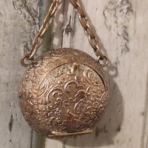 May include: A small, round, antique-style pendant with an ornate floral pattern. The pendant is made of a bronze-colored metal and hangs from a matching chain. The pendant is approximately 2 inches in diameter.