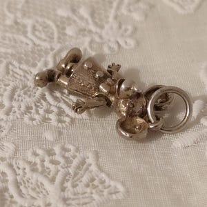 May include: A silver-toned charm in the shape of a small figure, possibly a doll or a person. The charm has a detailed design with visible facial features, clothing, and a ring for attachment. The charm is lying on a white, textured surface.