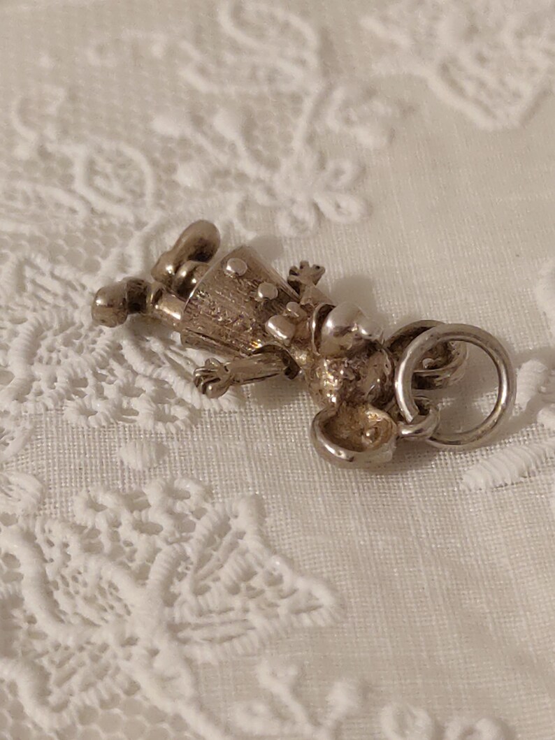 May include: A silver-toned charm in the shape of a small figure, possibly a doll or a person. The charm has a detailed design with visible facial features, clothing, and a ring for attachment. The charm is lying on a white, textured surface.