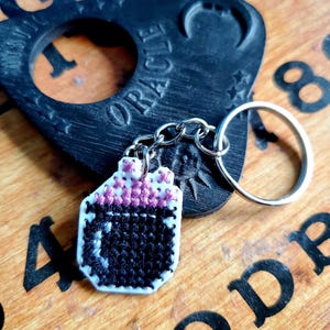 May include: A black and white cross-stitch keychain charm shaped like a cauldron with a pink rim and stars. The charm is attached to a silver chain and a silver key ring.