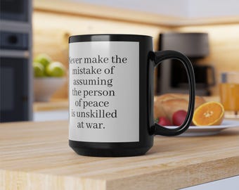 Puede incluir: Taza de café negra con interior blanco y asa negra. La taza presenta la cita: "Nunca cometas el error de asumir que la persona de paz no está capacitada para la guerra". La taza está sobre una superficie de madera.