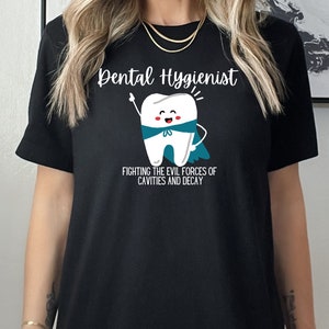 Puede incluir: Camiseta negra con un diente de dibujos animados blanco que lleva una capa azul y el texto "Higienista dental Luchando contra las fuerzas del mal de las caries y la descomposición".