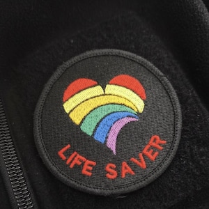 Könnte beinhalten: Schwarzer, gestickter Aufnäher mit einem Regenbogenherz-Design und dem roten Schriftzug "LIFE SAVER". Der Aufnäher ist rund und auf einem schwarzen Stoff befestigt.