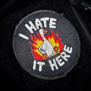 Könnte beinhalten: Ein runder, schwarzer Aufnäher mit dem weißen Schriftzug "I HATE IT HERE". In der Mitte befindet sich eine weiße Gans in Flammen in Rot, Orange und Gelb. Der Aufnäher ist auf einem schwarzen Stoff befestigt.