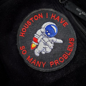 Könnte beinhalten: Runder, gestickter Aufnäher mit dem Text "HOUSTON I HAVE SO MANY PROBLEMS" in Rot. Der Aufnäher zeigt einen Astronauten mit blauem Helm und weißem Raumanzug auf schwarzem Hintergrund.