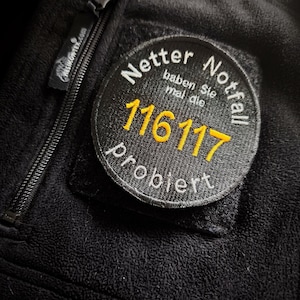 Könnte beinhalten: Nahaufnahme einer schwarzen Fleecejacke mit Reißverschluss und rundem Aufnäher. Der Aufnäher zeigt den Text "Netter Notfall" und "116117" in Gelb, mit zusätzlichem Text in Weiß. Die Jacke hat einen Helikon-Tex-Reißverschluss.