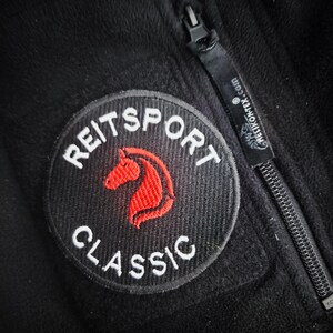 Könnte beinhalten: Schwarze Fleecejacke mit einem runden Aufnäher mit den weißen Worten "REITSPORT CLASSIC" und einem roten Pferdekopf. Ein schwarzer Reißverschluss und der Text "HELIKON-TEX.com" sind ebenfalls zu sehen.