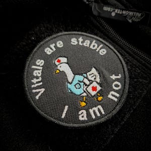 Könnte beinhalten: Ein runder, schwarzer Aufnäher mit einer weißen Gans als Krankenschwester, die ein blaues Oberteil und eine medizinische Tasche mit rotem Kreuz trägt. Der Text "Vitals are not stable I am" ist in Weiß um die Gans herum gestickt.