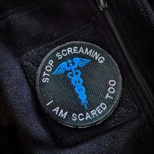 Könnte beinhalten: Schwarze Fleecejacke mit einem runden Aufnäher. Der Aufnäher zeigt ein blaues Caduceus-Symbol und den Text "STOP SCREAMING I AM SCARED TOO" in weißen Buchstaben. Ein schwarzer Reißverschluss ist sichtbar.