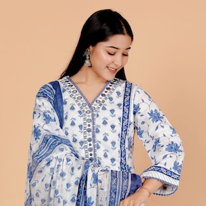 Könnte beinhalten: Weißes und blaues Kleid mit Blumenmuster und V-Ausschnitt. Das Kleid hat ein blaues Blumenmuster und blaue Verzierungen an den Ärmeln und am Ausschnitt. Es hat einen passenden Schal.