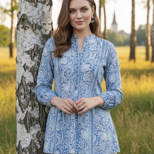 May include: A light blue tunic with a white floral pattern. The long-sleeved top has a mandarin collar and a button-down front. The tunic is paired with white pants. The background features a birch tree and a field of tall grass.