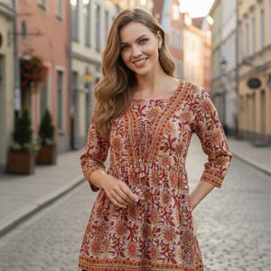 May include: A three-quarter sleeve tunic featuring a light pink base with a repeating floral pattern in shades of red, orange, and yellow. The garment has a round neckline, a gathered waistline, and a decorative front placket with tassels.