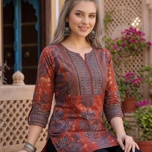 May include: A rust-colored tunic featuring a dark blue floral and leaf print, a V-neckline with vertical patterned detailing, and three-quarter length sleeves with matching cuffs. The garment is paired with solid black pants and silver jewelry.