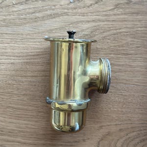 Op de afbeelding: Een T-vormig sanitair koppelstuk van gepolijst messing met een cilindrisch lichaam, een zijdelingse uitlaat met schroefdraad en een platte bovenflens die is vastgezet met een kleine centrale schroef. Het metalen oppervlak vertoont slijtage.