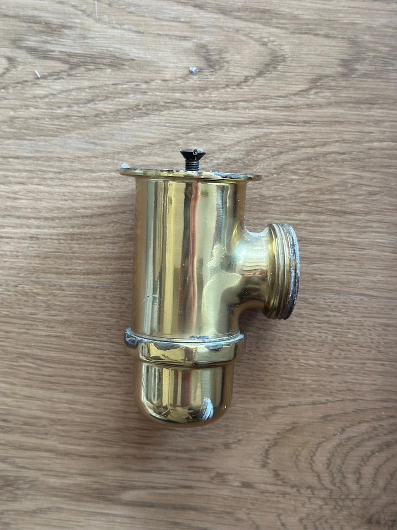 Op de afbeelding: Een T-vormig sanitair koppelstuk van gepolijst messing met een cilindrisch lichaam, een zijdelingse uitlaat met schroefdraad en een platte bovenflens die is vastgezet met een kleine centrale schroef. Het metalen oppervlak vertoont slijtage.