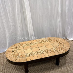 Peut inclure: Une table basse ovale avec un cadre en bois brun foncé et quatre pieds courts. Le plateau présente une grille de carreaux carrés beige clair au centre, entourée d'une bordure de carreaux rectangulaires ornés d'imprimés botaniques brun foncé.