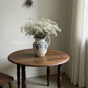 Puede incluir: Una jarra de cerámica con un patrón floral azul llena de flores blancas de paniculata. La jarra cuenta con un asa curva y un pico, colocada sobre una mesa redonda de madera con patas torneadas. El texto Collectiondemaman es visible.