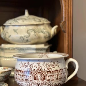 Peut inclure: Un pot de chambre en céramique blanche avec un motif floral brun et un portrait de femme dans un médaillon. Le pot a une seule poignée et un corps arrondi.