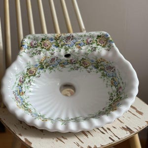 Może przedstawiać: Biała ceramiczna umywalka z falistym brzegiem i kwiatowym wzorem w żółte, niebieskie i zielone kwiaty. Umywalka posiada centralny otwór odpływowy oraz mniejszy otwór przelewowy przy górnej krawędzi, ozdobiony ciągłą kwiatową bordiurą.