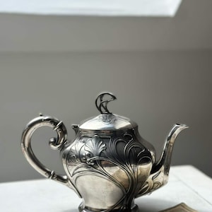 Op de afbeelding: Een zilverkleurige metalen theepot met een gedetailleerd, reliëf bloemen- en bladpatroon op de behuizing. De theepot heeft een gebogen handvat, een taps toelopende tuit en een deksel met een decoratieve knop. Het oppervlak is gepolijst.