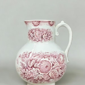 May include: White ceramic jug with a rounded body and a curved handle. It features a repeating floral pattern in pink and red hues. The floral design includes roses and other flowers, creating a vintage aesthetic.