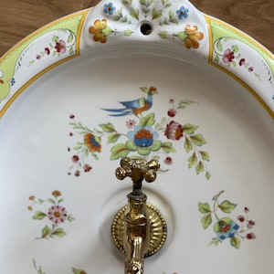 Op de afbeelding: Een ovale witte keramische wandwastafel met een koperen kraan en kleurrijke handgeschilderde bloem- en vogelmotieven. De rand van de wastafel is versierd met gele en groene randen, terwijl het midden gedetailleerde illustraties van vogels toont.