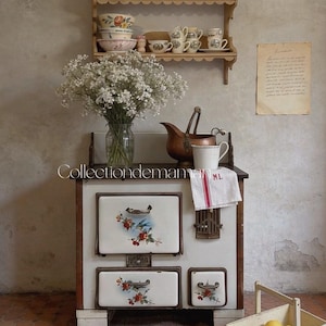 Könnte beinhalten: Ein weißer Emailleherd mit Blumenmotiven, ausgestattet mit zwei Ofentüren und einem Seitenfach. Oben stehen eine Glasvase mit weißen Blumen, ein Kupferkrug und eine weiße Tasse mit einem Geschirrtuch, das mit ML gekennzeichnet ist.