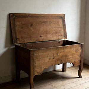 May include: A rectangular wooden chest with a hinged lid in an open position. The chest features a natural brown wood grain finish, a flat top panel, and four legs with curved, scalloped apron detailing along the front and side base edges.