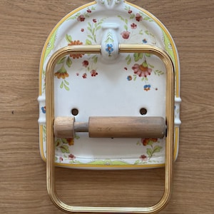 Op de afbeelding: Een wandgemonteerde toiletrolhouder met een witte keramische achterplaat voorzien van een bloemenpatroon in rood, oranje, groen en blauw. Het armatuur bevat een goudkleurig metalen rechthoekig frame en een cilindrische houten rol.