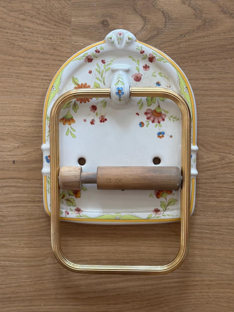 Op de afbeelding: Een wandgemonteerde toiletrolhouder met een witte keramische achterplaat voorzien van een bloemenpatroon in rood, oranje, groen en blauw. Het armatuur bevat een goudkleurig metalen rechthoekig frame en een cilindrische houten rol.