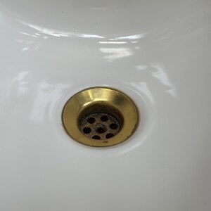 Op de afbeelding: Een witte keramische wastafel met een ronde, messingkleurige metalen afvoer. De afvoer bevindt zich in het midden van de wastafel en bevat een geperforeerde zeef met meerdere kleine gaatjes, met een metalen afwerking die lichte gebruikssporen vertoont.