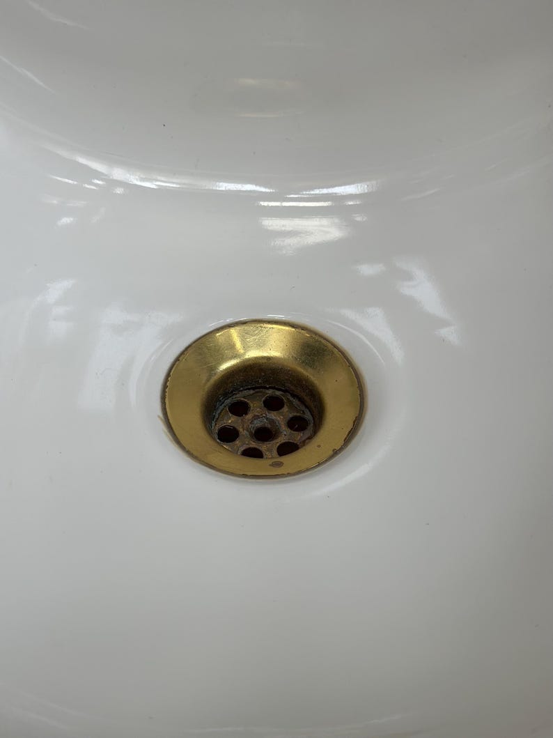 Op de afbeelding: Een witte keramische wastafel met een ronde, messingkleurige metalen afvoer. De afvoer bevindt zich in het midden van de wastafel en bevat een geperforeerde zeef met meerdere kleine gaatjes, met een metalen afwerking die lichte gebruikssporen vertoont.