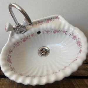 Könnte beinhalten: Ein weißes Keramikwaschbecken in Muschelform mit einem zarten rosa Blumenmuster am Rand. Das Becken verfügt über eine Chromarmatur mit weißen Porzellangriffen und einen mittigen Chrom-Abfluss mit einem perforierten Gitterdesign.