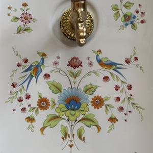 Op de afbeelding: Een witte keramische wandwastafel met een koperen kraan en een kleurrijk bloemenpatroon met twee vogels. Het ontwerp bevat blauwe, groene, oranje en roze bloemen, groene bladeren en twee vogels met blauw en oranje verenkleed aan weerszijden.