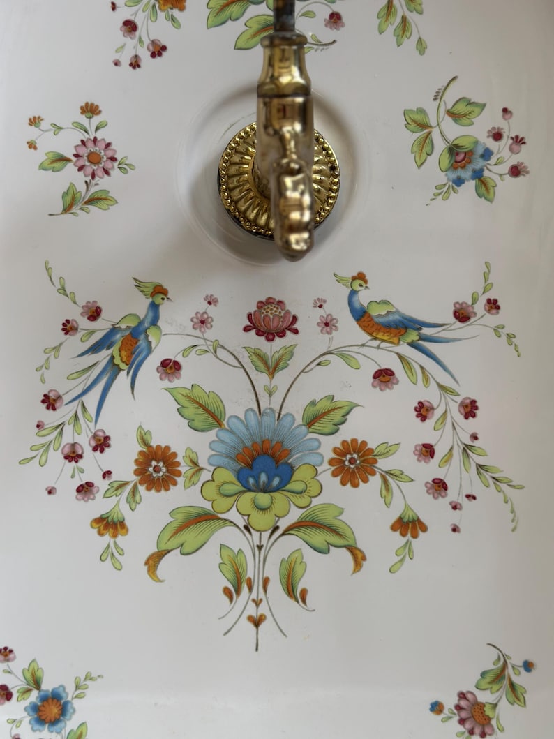 Op de afbeelding: Een witte keramische wandwastafel met een koperen kraan en een kleurrijk bloemenpatroon met twee vogels. Het ontwerp bevat blauwe, groene, oranje en roze bloemen, groene bladeren en twee vogels met blauw en oranje verenkleed aan weerszijden.