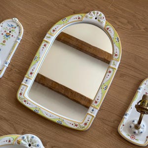 Op de afbeelding: Een boogvormige keramische wandspiegel met een wit frame voorzien van handgeschilderde bloemmotieven in groen, rood en blauw. De spiegel heeft een decoratieve bovenrand met een gat voor montage en een glad, reflecterend glazen oppervlak.