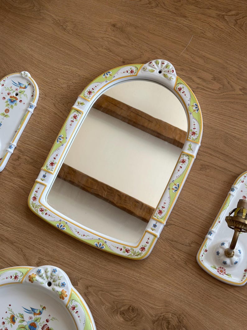 Op de afbeelding: Een boogvormige keramische wandspiegel met een wit frame voorzien van handgeschilderde bloemmotieven in groen, rood en blauw. De spiegel heeft een decoratieve bovenrand met een gat voor montage en een glad, reflecterend glazen oppervlak.