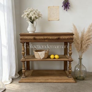Peut inclure: Une console en bois brun clair à deux niveaux, dotée de détails sculptés sur le bord supérieur et de pieds tournés. La table supporte un pichet blanc avec des fleurs blanches, un panier en osier avec un tissu et deux citrons jaunes sur l'étagère.