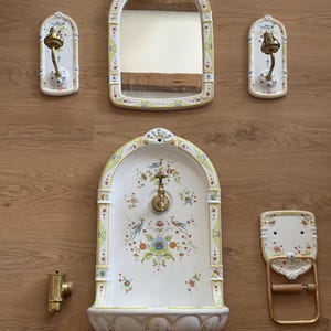 May include: A ceramic bathroom set featuring a wall-mounted fountain with a brass faucet, an arched mirror, two wall sconces, a towel ring, and a separate brass valve. Each piece is white with yellow trim and a colourful floral and bird pattern.