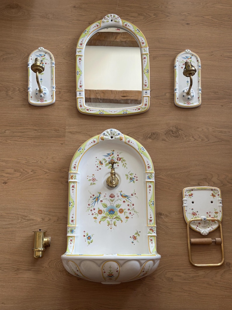 Op de afbeelding: Een keramische badkamerset bestaande uit een wandfontein met een messing kraan, een boogvormige spiegel, twee wandlampen, een handdoekring en een losse messing klep. Elk item is wit met een gele rand en een kleurrijk bloemen- en vogelpatroon.