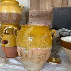 Peut inclure: Un pot en terre cuite rustique présentant une partie supérieure émaillée jaune vif et une base en terre cuite naturelle non émaillée. Le récipient possède deux petites anses latérales, une large ouverture et une surface patinée.