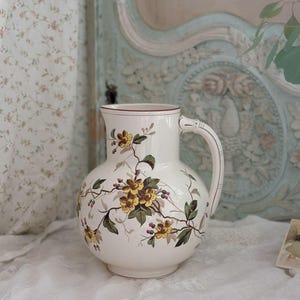 Peut inclure: Pichet en céramique blanche avec un corps arrondi et une anse. Il est orné d'un motif floral avec des fleurs jaunes et marron, des feuilles vertes et des baies rouges. Le bord est bordé de marron.
