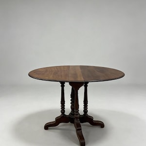 May include: A round wooden drop-leaf table with a dark brown finish. The table features a smooth circular top that can be folded down and is supported by a central pedestal base with four curved legs and decorative turned spindle details.